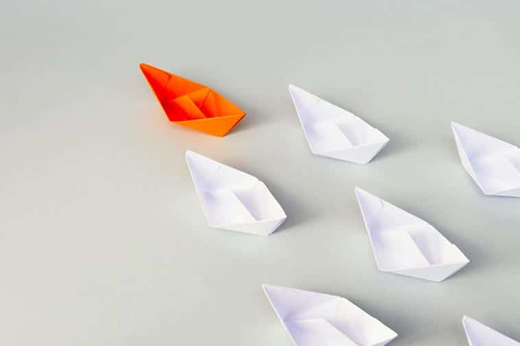 an orange paper boat leading six other white boats behind